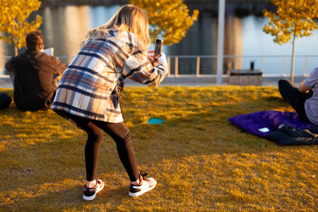 Back view of young woman taking selfie with mobile phone in autumn parkの写真素材