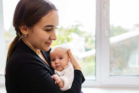 Young mother holding her newborn baby boy in her arms, looking out the window