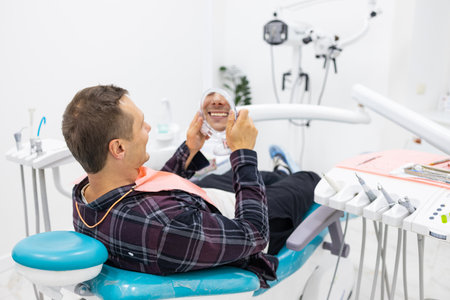 Happy Handsome Caucasian Man Looking To Mirror After Teeth Treatment In Modern Clinic, Male Patient...