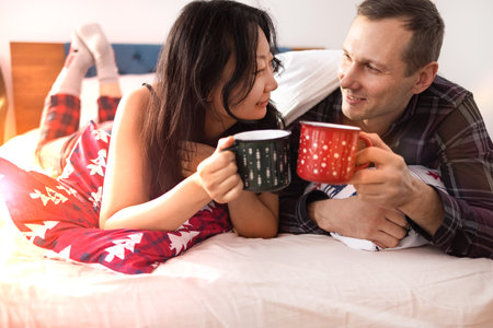 Couple in love lying in bed and drinking coffee with Christmas cups. Man and woman in pajamas.の写真素材