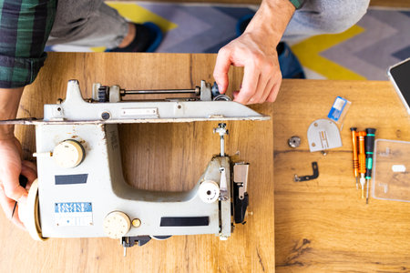 Repairman master is testing old disassembles sewing machine in workshop repairing it sitting at table. Man is looking inside sewing machine trying to repair it, hands closeup.の写真素材