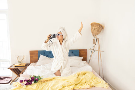 Young woman in a white bathrobe and with a towel on her head is taking pictures with a retro camera.の写真素材