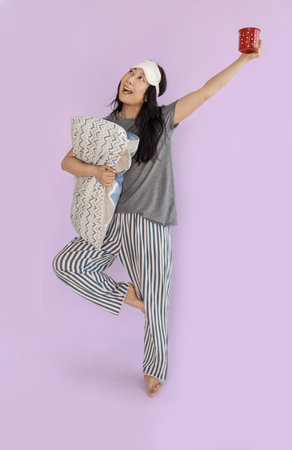 Full body smiling happy young Asian woman wears pyjamas jam sleep eye mask rest relax at home hold...