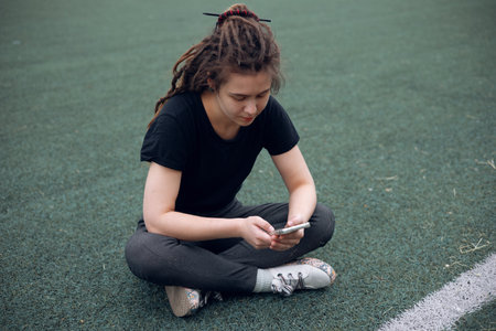 Lonely young Caucasian girl using smartphone while sitting outdoors near playground.の写真素材