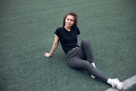 A happy stylish young teenager with long dreadlocks sits on the grass on the playground. Daytime, stadium, open airの写真素材