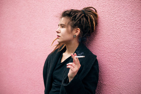 Portrait of a young girl with dreadlocks in a black coat smoking a cigarette on a pink backgroundの写真素材