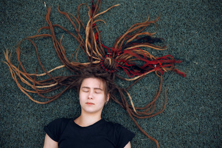 A hipster teen girl with dreadlocks lies on the grass on the playground. Daytime, stadium, open air.の写真素材