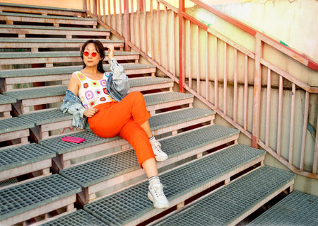 Young hipster Asian girl in orange pants and a knitted top sitting on metallic stairs relaxing in the city. Street fashion concept.の写真素材