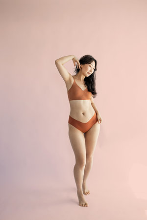 Full length portrait of a beautiful Asian woman in orange bikini posing isolated over pink backgroundの写真素材