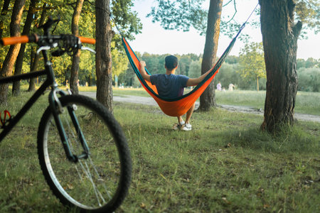 Biker man sitting in orange hammock in pine forest. Biking and camping concept.の写真素材