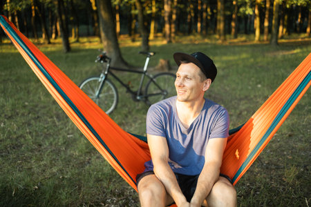 Young man travels on bicycle, relaxing in red hammock, rest in forest near lake. Happy cyclist in hammock at campsite by river. Male on bike in hammock at sunset.の写真素材