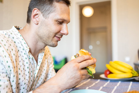 Hungry young Caucasian man eating burger in kitchen at night.の写真素材
