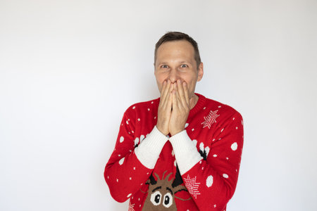 Caucasian young man in Christmas sweater covering his mouth and looking at camera over white backgroundの写真素材