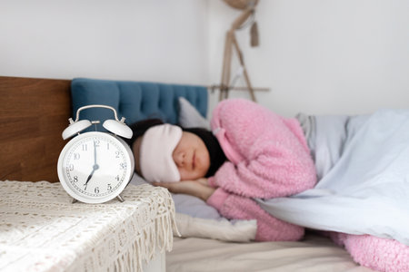 white alarm clock set for 7 AM on bedside table in bedroom, with happy peaceful tranquil young woman sleeping on bed in blurred background. Sleep, morning, daily routine conceptsの写真素材