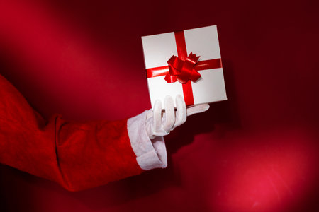 Santa Claus hand holding a gift box with red ribbon on red backgroundの写真素材