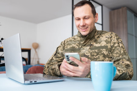 Portrait of a smiling soldier using his mobile phone and laptop at homeの写真素材