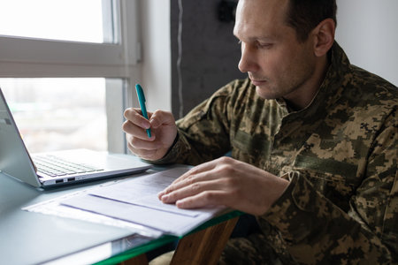 Soldier sitting at his desk in front of laptop and writing in notebookの写真素材