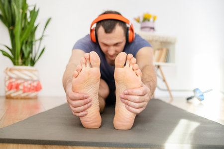 A Caucasian man with headphones stretching his legs on yoga mat at home.