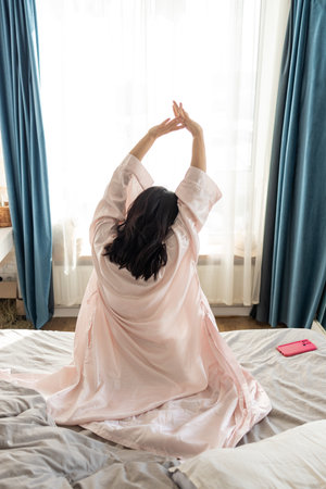 Happy Asian woman on bed awakened from enough and healthy sleep. Woman stretching in bed after wake up, back view.の写真素材