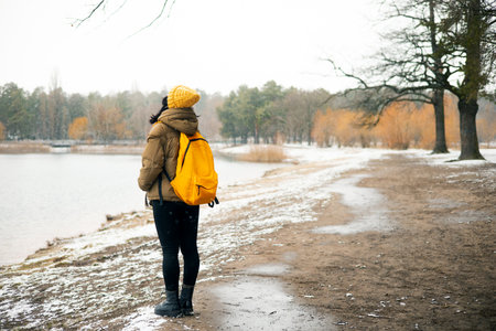 A girl with a yellow backpack walks along the bank of a frozen lake.の写真素材