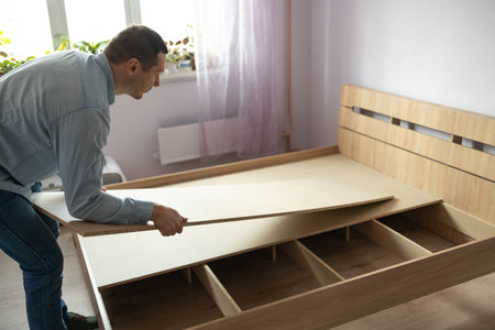 Assembling a New bed in the Bedroom. Man tightened the furniture on the bed frame. Self-assembly DIY using a guide. House design concept.の写真素材