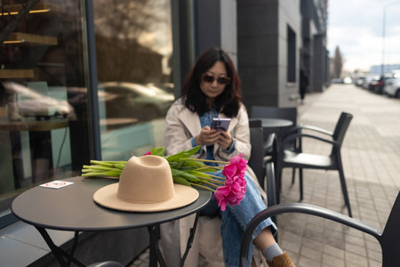 Stylish Asian woman using her phone for messaging, communication, content creating for social media sitting in outdoor cafe. Girl holding tulips bouquet.の写真素材