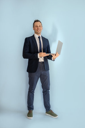 Full length portrait of mature man holding laptop while standing against blue backgroundの写真素材