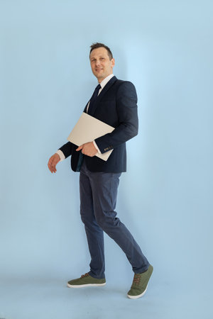 Full length portrait of a confident businessman holding laptop while standing against blue backgroundの写真素材