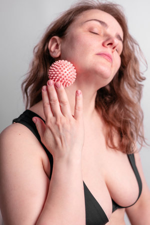 Portrait of pleased young brunette woman with closed eyes massaging neck with manual massage ball at home, lymphatic system support and home-based massage, balancing energyの写真素材