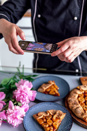 happy pastry chef woman taking photo of fruit pie on cellphone at cozy kitchenの写真素材