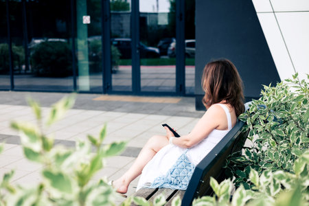 Young woman sitting on a bench and using a mobile phone outside near building. Lifestyle technology modern sustainable architecture concept.の写真素材