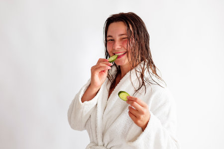 Close up portrait of happy young beautiful woman holding sliced cucumber smiling while looking at camera and eating. Caucasian positive female in robe on white background.の写真素材