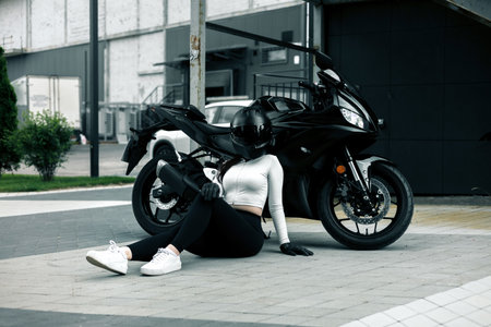 Biker girl in sportswear sitting on the ground near her motorcycle.