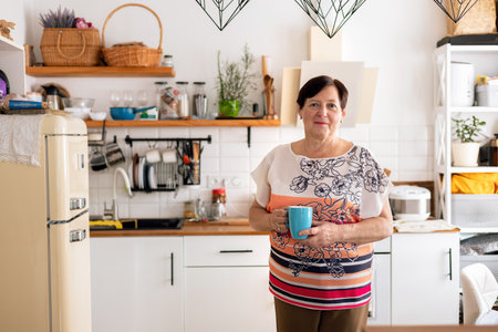 Portrait of happy senior woman standing in kitchen with cup of coffeeの写真素材