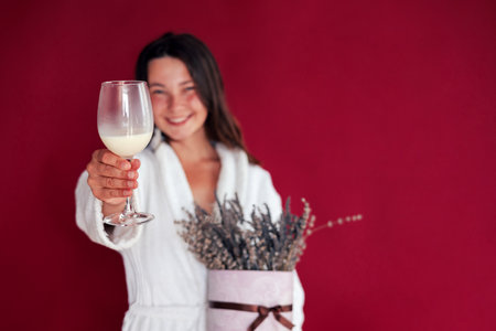 Happy woman in bathrobe holding glass of tasty dairy drink on red background.の写真素材