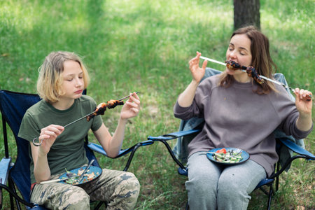 Mother and son are sitting on a camping chair in the park and eating barbecueの写真素材
