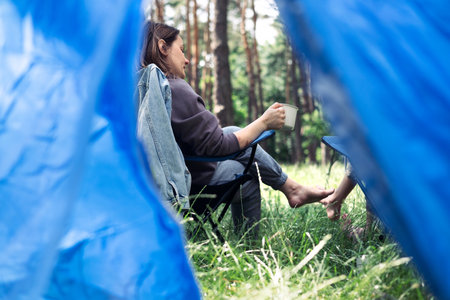 Woman camping in the forest, she is sitting near a tent and drinking coffeeの写真素材