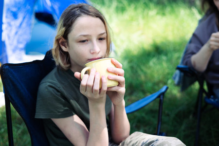 Teenage boy sitting in camping chair and drinking tea from a cupの写真素材