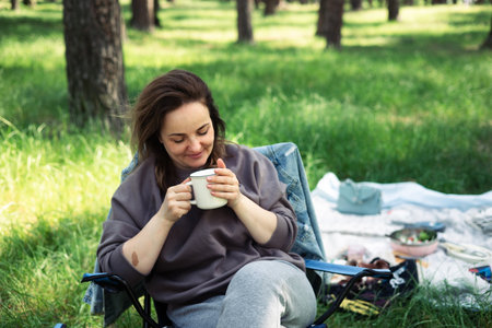 Young woman sitting on a folding chair in the park and drinking coffeeの写真素材