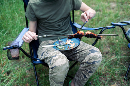 A man in camouflage sits on a camping chair and eats grilled meat.の写真素材