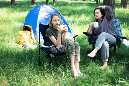 Mother and daughter drinking tea and eating sandwich on camping chair in parkの写真素材