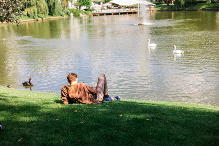 Young man sitting on the grass by the lake in city park, back view.の写真素材