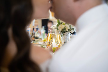 Bride and groom kissing and holding wedding champagne glasses while sitting at restaurant table on...