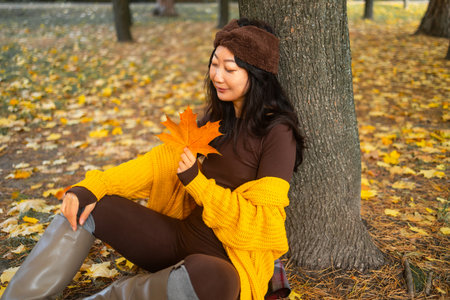 Beautiful young woman sitting on the ground in autumn park and holding a maple leafの写真素材