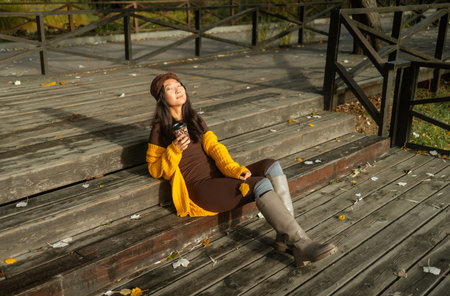 Young Asian woman in yellow sweater, scarf and boots sitting on wooden bridge and drinking coffee in autumn park.の写真素材