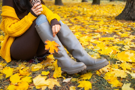 A young woman in a yellow sweater and boots sits on the ground in the autumn park.の写真素材