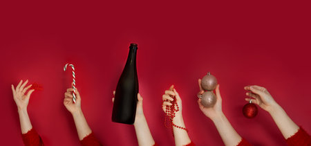 Female hands holding Christmas balls, candy cane, bottle of champagne and gift box on red...