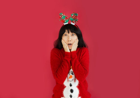 Asian woman wearing Santa hat and christmas sweater against a red background, looking surprised with...