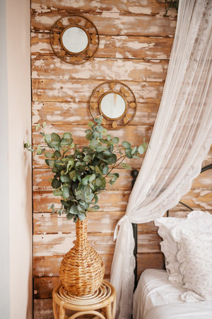 Details of bright elegant interior in cozy bedroom. Wicker side table with vase and flowers next to comfortable bed, two roung mirror on wall.の写真素材