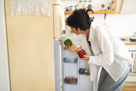 Side view of young woman looking at fresh vegetables in refrigerator at homeの写真素材